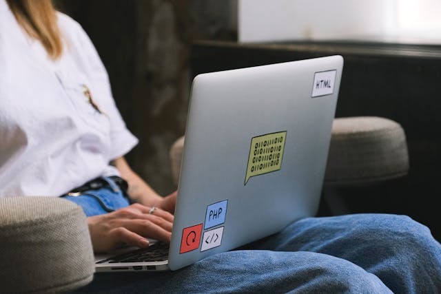 A woman sitting on a chair, focused on her laptop, with a cozy room in the background.