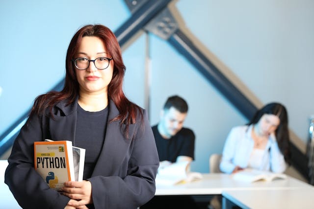 A woman in glasses holds a book, engaging a group of attentive students in a classroom setting.
