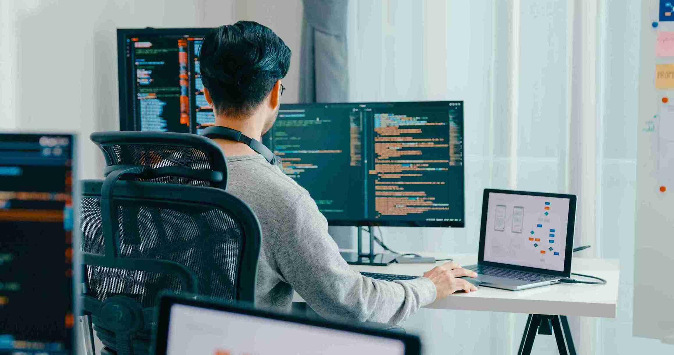 A man sits at a desk with two computers, focused on his work in a modern office setting.